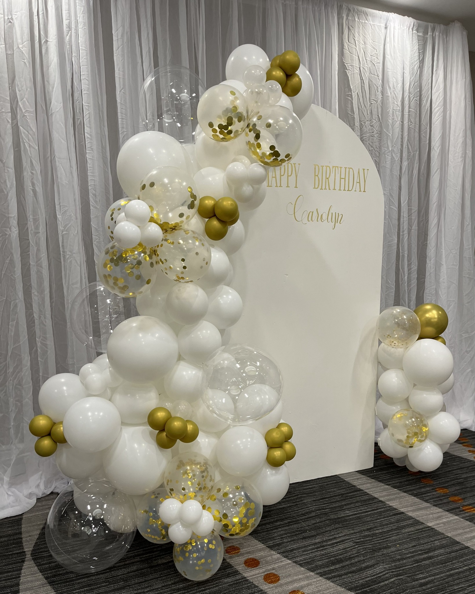 A personalized white birthday welcome sign framed by white, clear and gold balloons against ivory draping