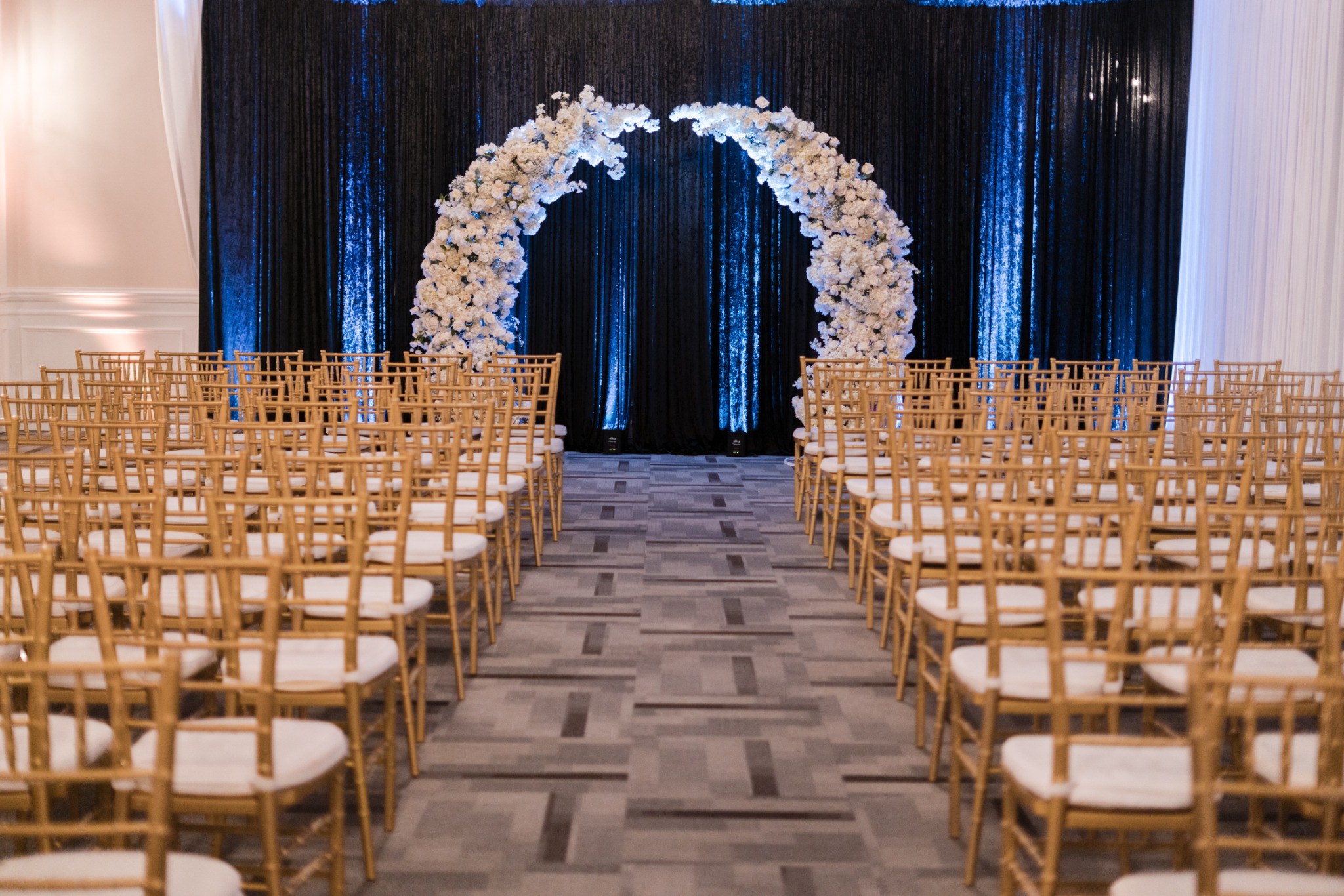 Gold ceremony chairs leading to a white floral arch against black draping