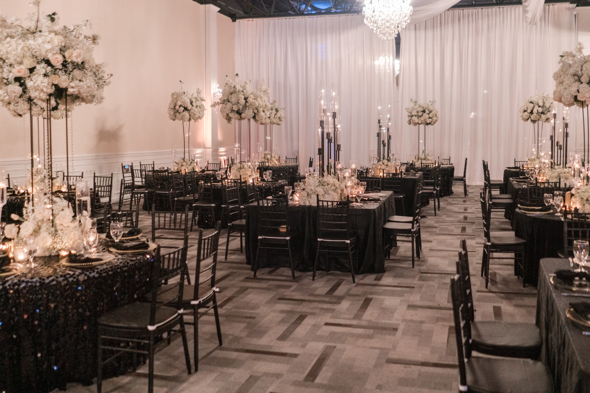 A reception room with black linens, tall white floral centerpieces, chandeliers and ivory draping