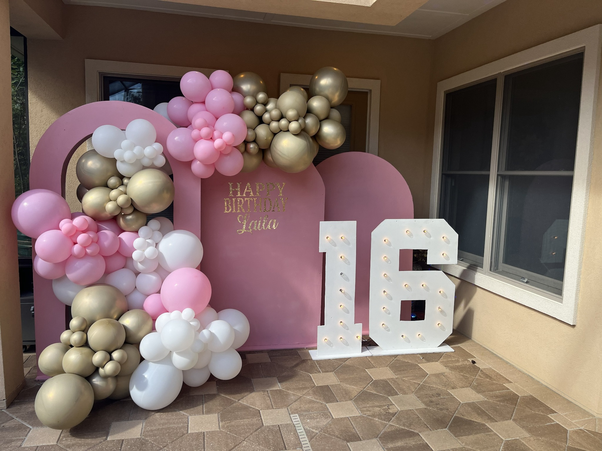 A pink birthday backdrop with pink, white and gold balloons and illuminated number sixteen letters